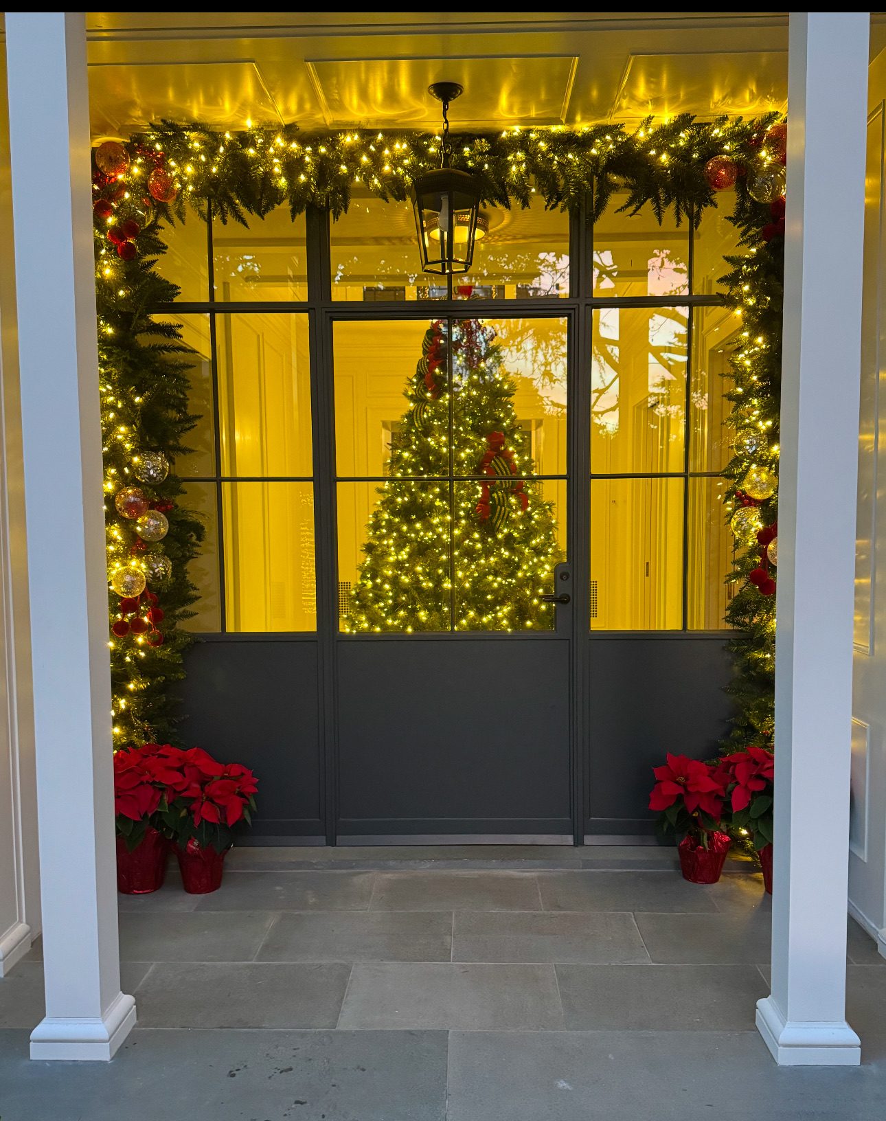 A front entry with lit garland and a glowing tree inside the glass door